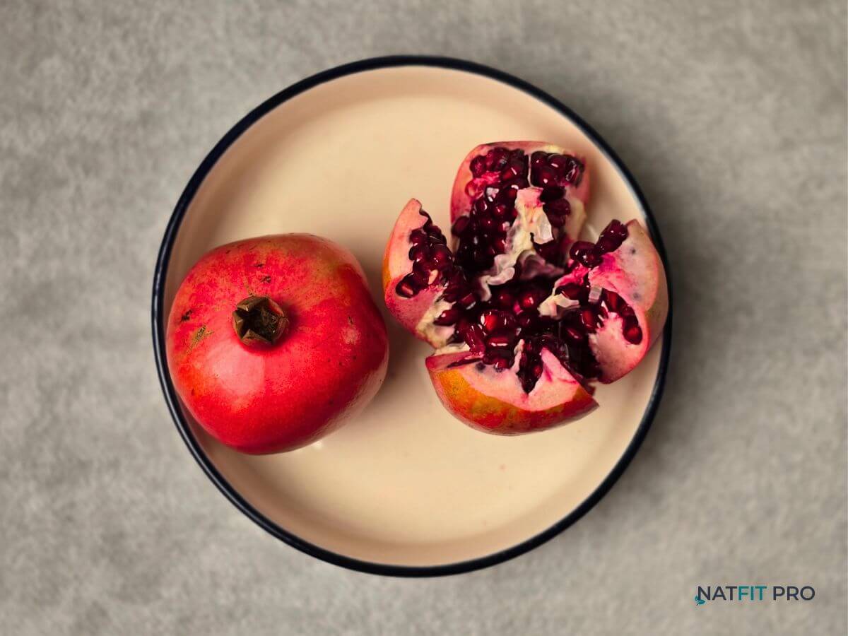 A whole pomegranate and a sliced open pomegranate revealing its ruby-red seeds, displayed on a cream plate - known for supporting blood flow with natural nitrates and antioxidants.