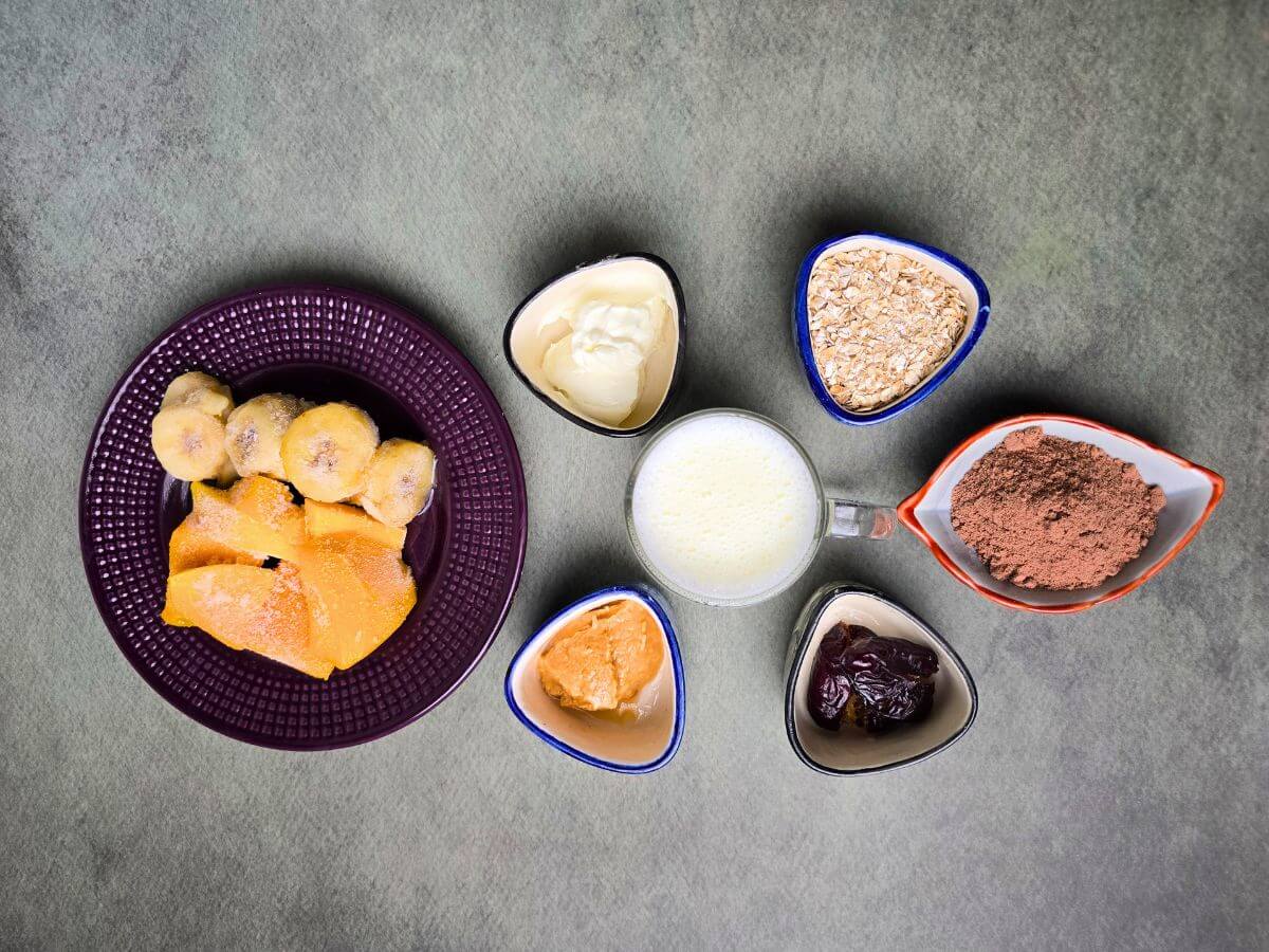 A flat lay of the ingredients for a muscle-gain smoothie, including frozen banana, mango, oats, dates, peanut butter, Greek yogurt, and chocolate whey protein powder.