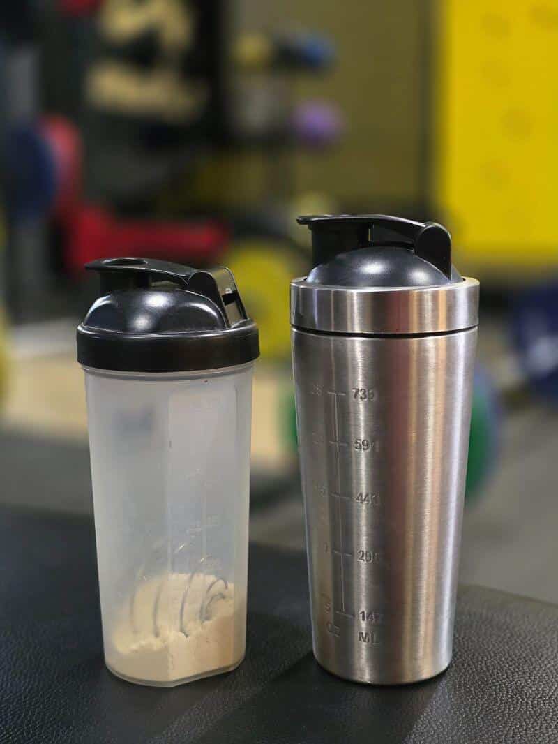 Two gym shaker bottles side by side, one metal and one plastic with protein powder inside