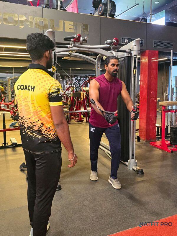 A client (Tamilarasan) preparing for a cable fly exercise while his personal trainer observes his setup in the gym.